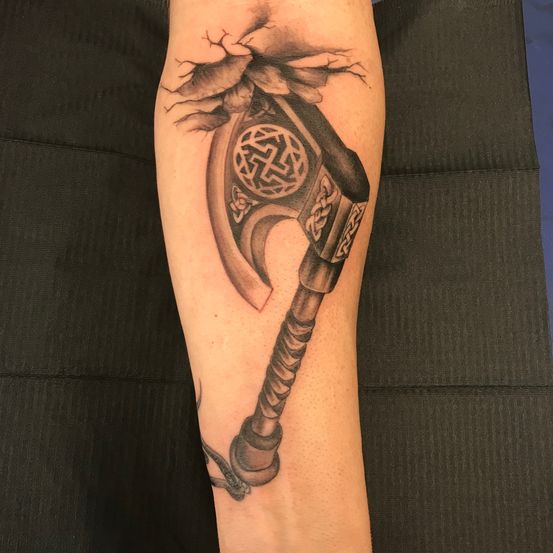 Ein tätowierter Arm zeigt ein detailliertes Design einer Axt, die durch die Haut bricht, mit einem keltischen Knoten auf der Klinge.