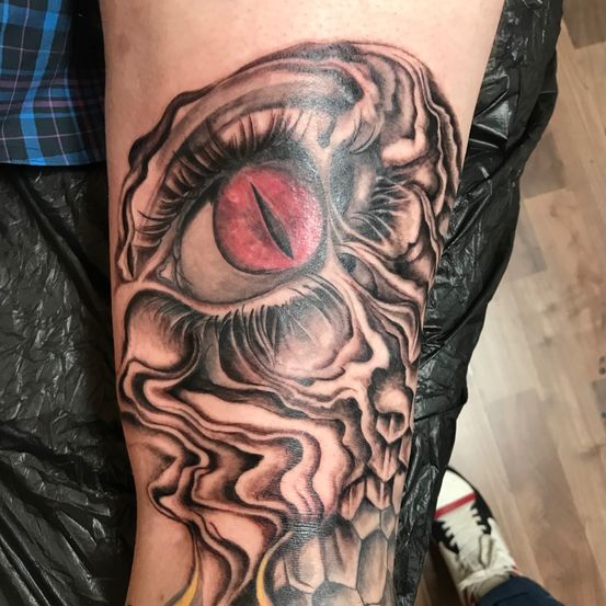 Ein detailliertes Tattoo eines Schädels mit einem roten Auge ist auf dem Arm einer Person zu sehen. Der Schädel hat eine komplexe Schattierung und ein Flammendesign unterhalb des Auges.