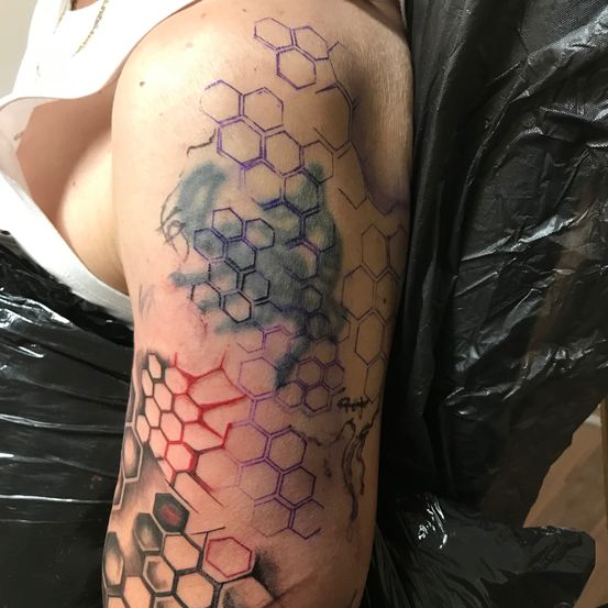 Der Arm einer Person mit einem großen Aquarell-Tattoo in Wabenform in verschiedenen Farben, darunter Lila, Blau und Rot, sitzt in einer schwarzen Plastiktüte.