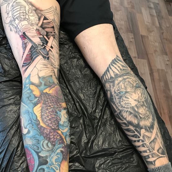 Eine Person mit Tätowierungen an beiden Beinen, eines zeigt einen Samurai mit einem Schwert und das andere einen Tiger, liegt auf einem schwarzen Laken.