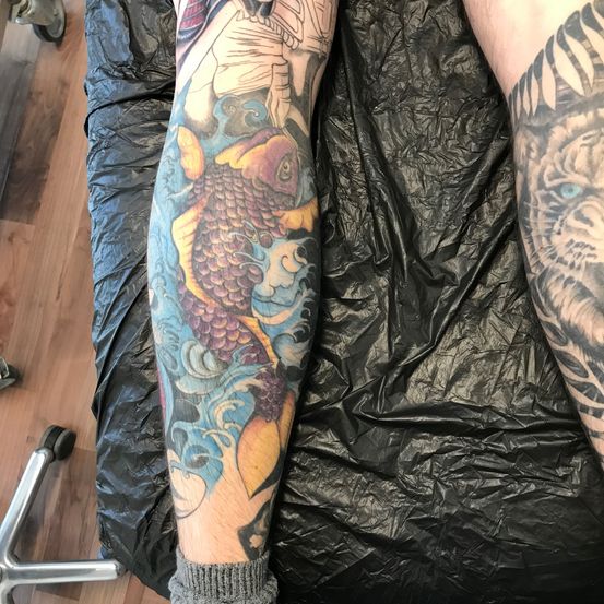 Eine Nahaufnahme eines Beins mit einem detaillierten Tattoo eines Fisches in Blau- und Lilatönen auf einem Holzboden, bedeckt mit einem schwarzen Laken.