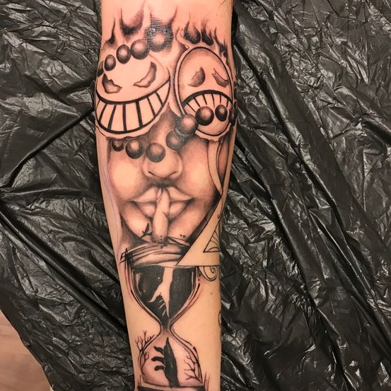 Ein Tattoo mit zwei grinsenden Gesichtern, einem Finger über dem Mund, vor einer Sanduhr und einem Dreieck, auf dem Arm einer Person.
