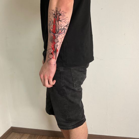 Ein Mann mit einem schwarzen Shirt und Shorts steht mit dem Rücken zu einer weißen Wand und zeigt ein detailliertes Tattoo an seinem linken Arm. Das Tattoo zeigt einen Baum mit einem roten und schwarzen Farbschema und eine DNA-Struktur, die sich mit den Ästen verflicht.