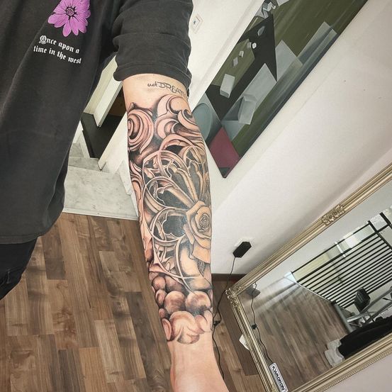Eine Person mit einem schwarzen T-Shirt zeigt ein Tattoo auf dem rechten Arm. Das Tattoo zeigt komplizierte Designs, darunter Wirbel und eine Rose, vor einem wolkenverhangenen Hintergrund. Hinter ihm hat eine Wand ein großes abstraktes Gemälde und ein Spiegel reflektiert eine Treppe.