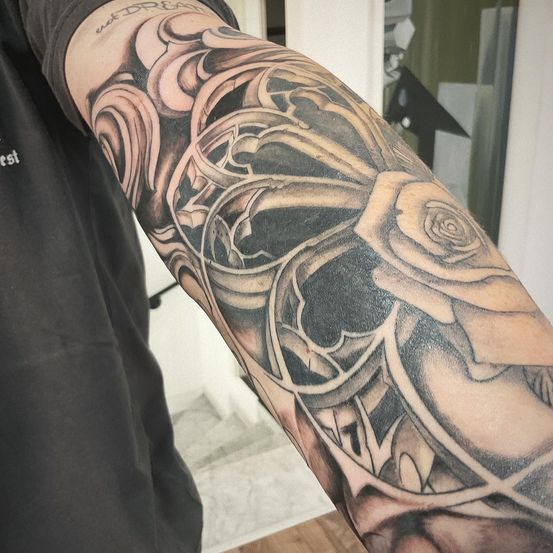 Eine Person in einem schwarzen T-Shirt zeigt ein detailliertes Tattoo am Arm. Das Tattoo zeigt eine Rose mit komplizierten Designs und Schattierungen.