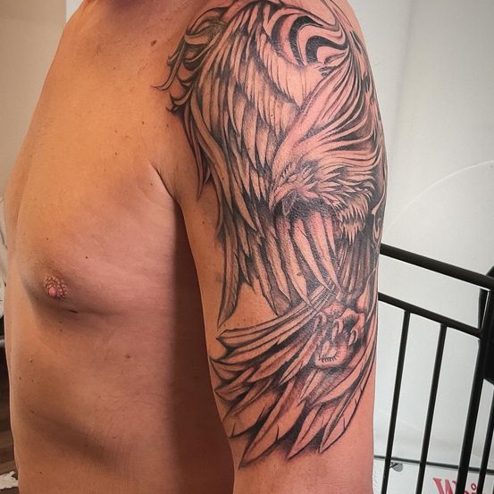 Der Oberarm eines Mannes zeigt ein großes, detailliertes Tattoo eines Vogelflügels, mit kunstvollen Mustern schattiert. Das Tattoo erstreckt sich vom Schulterbereich bis zum Ellbogen.