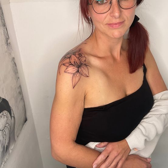 Eine Frau mit roten Haaren und Brille posiert für ein Foto mit einem Blumentattoo auf ihrer Schulter. Sie trägt ein schwarzes Oberteil und hat ihren Arm um jemanden.