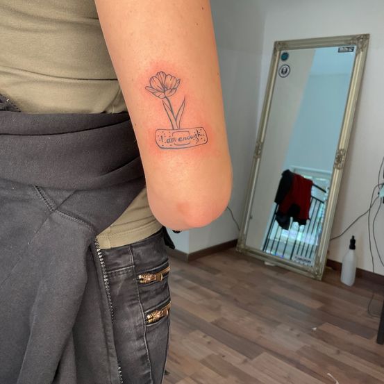 Ein Arm eines Menschen mit einem Blumentattoo, das in einem kleinen Topf 'Ich bin genug' steht. Die Person trägt ein graues Shirt und Hose.
