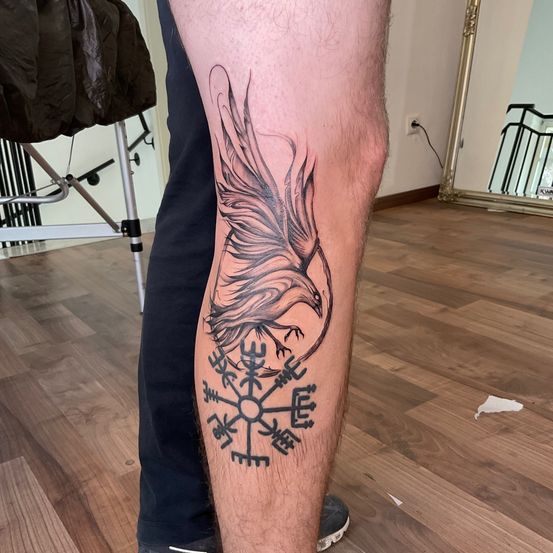 Ein Beintattoo eines Wikingeradlers mit einem komplizierten Runensymbol darunter. Das Tattoo ist detailliert mit Schattierungen und schwarzen Linien.