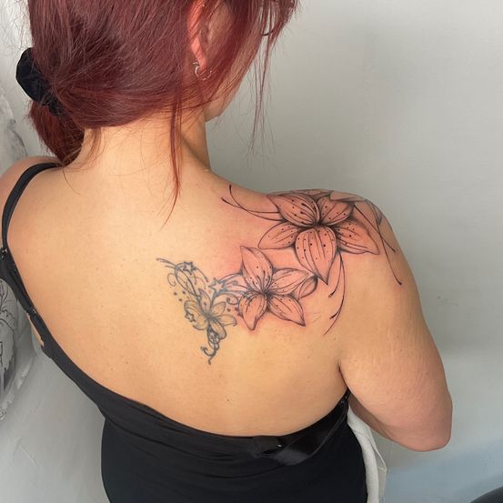 Eine Frau mit roten Haaren zeigt ein Tattoo auf ihrer Schulter, das ein detailliertes Design von Blumen und einem Schmetterling zeigt.