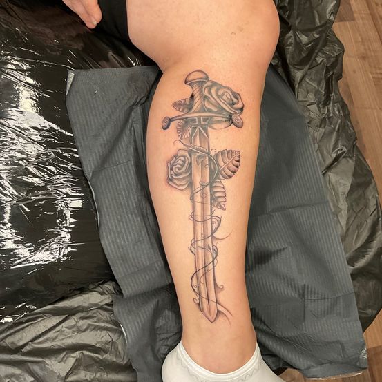 Ein Tattoo eines Schwertes mit Rosen und Blättern auf dem Bein einer Person, auf einem grauen Tuch ausgestellt.