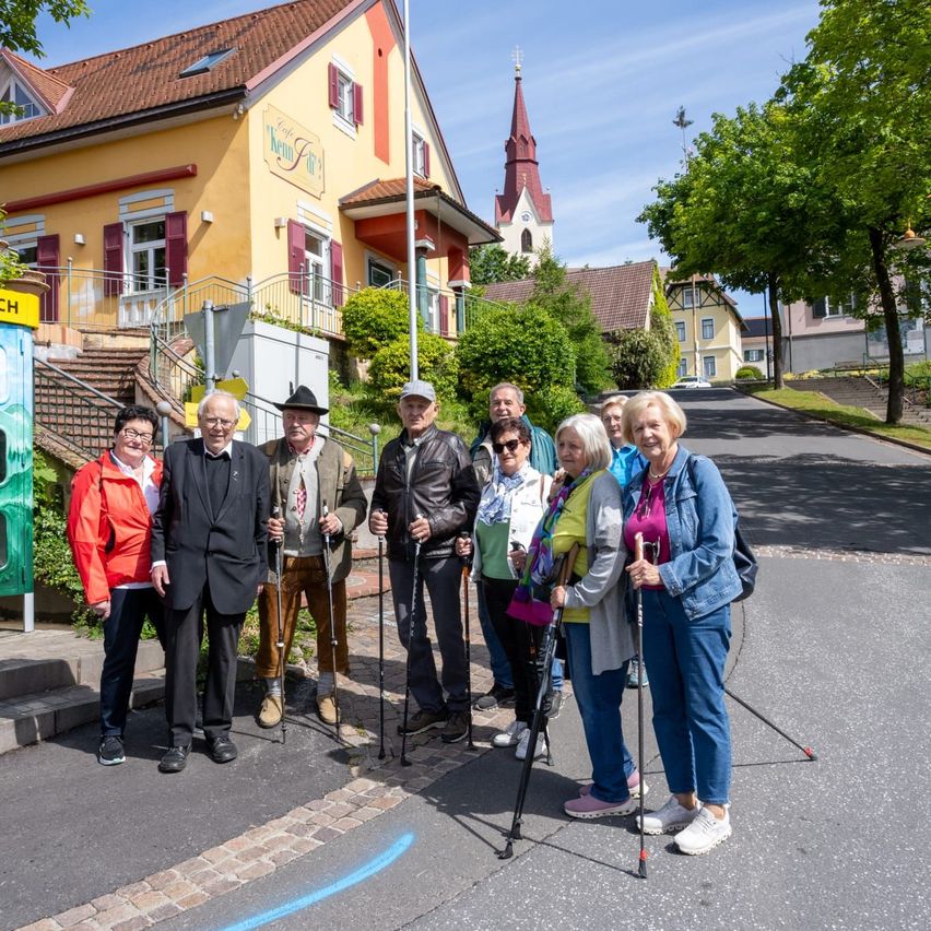 Eine Gruppe älterer Menschen steht mit Wanderstöcken auf einer Straße vor einem Gebäude mit rotem Dach und Kirchturm.