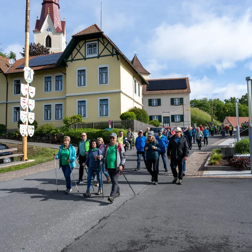 Eine Gruppe von Menschen geht auf einer Straße, die von Gebäuden auf beiden Seiten gesäumt ist. Sie tragen Jacken, Hüte und Brillen, und einige halten Trekkingstöcke. Das Gebäude auf der linken Seite hat Solarmodule auf seinem Dach.