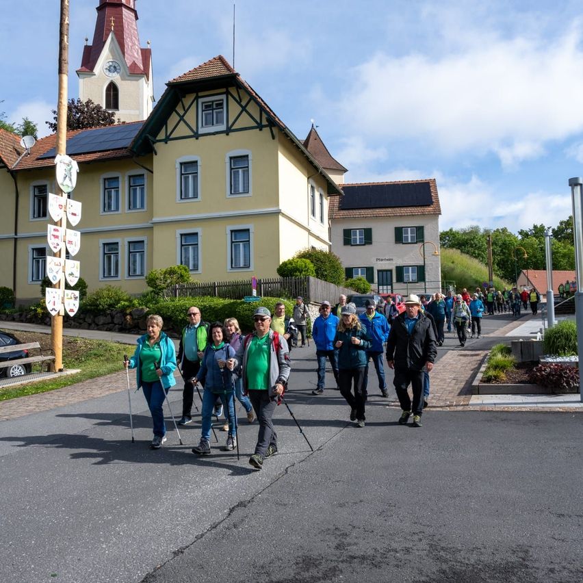 Eine Gruppe von Menschen geht auf einer Straße, die von Gebäuden auf beiden Seiten gesäumt ist. Sie tragen Jacken, Hüte und Brillen, und einige halten Trekkingstöcke. Das Gebäude auf der linken Seite hat Solarmodule auf seinem Dach.