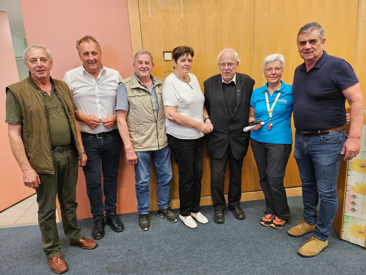 Eine Gruppe von Erwachsenen, wahrscheinlich Kollegen, steht zusammen für ein Foto. Sie sind lässig gekleidet und posieren vor einem hölzernen Hintergrund.