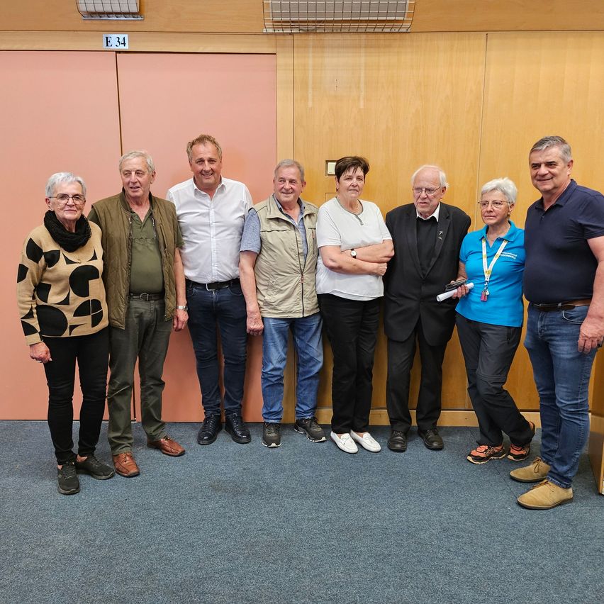 Eine Gruppe von acht Menschen, darunter ältere Erwachsene, posiert für ein Foto vor einer Holzwand mit einer rosa Tür, die mit E34 beschriftet ist. Sie stehen in einer Reihe mit einer Mischung aus Jacken und Anzügen.