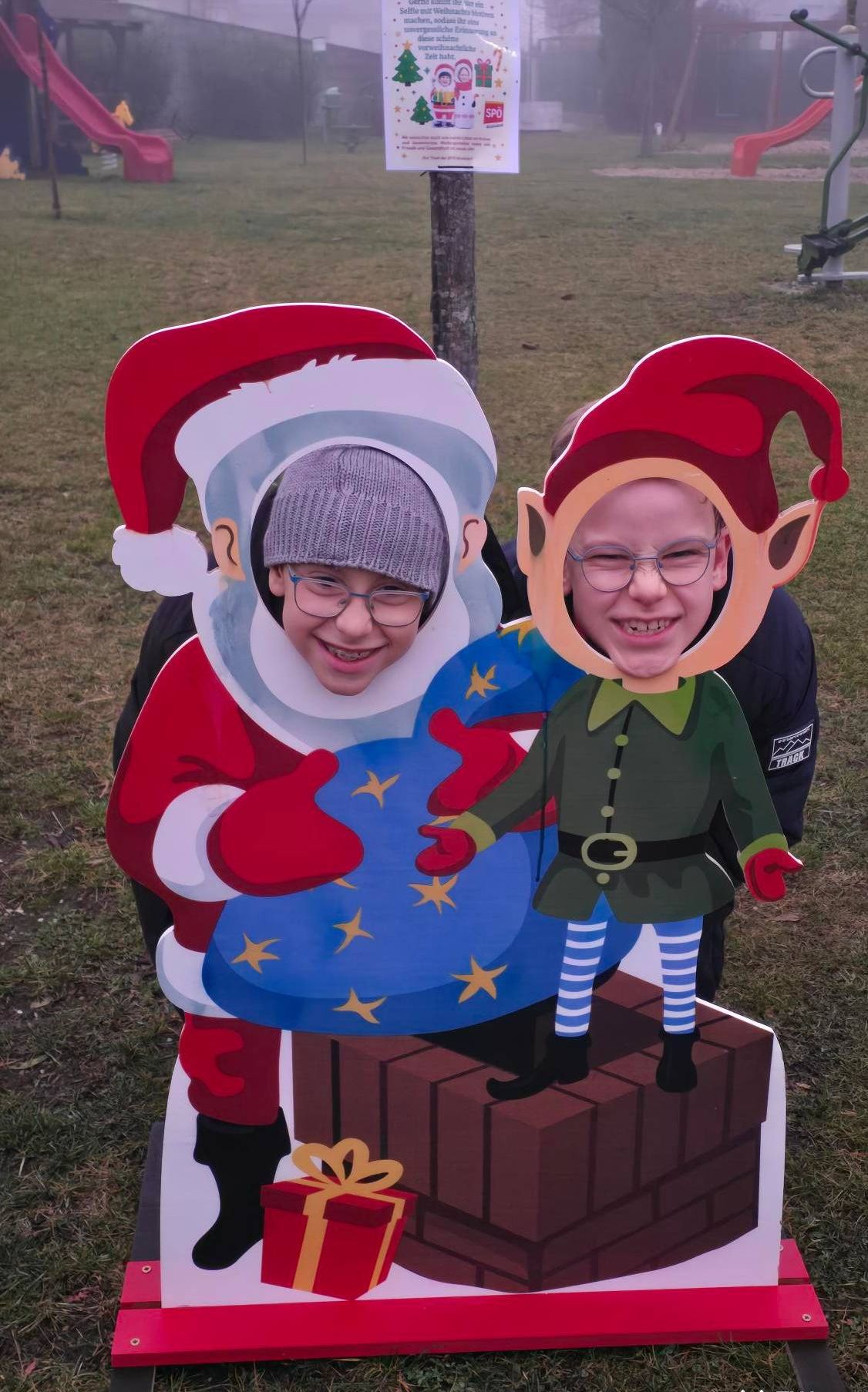 Zwei Kinder in Ausschnitten von Santa Claus und einem Elfen gekleidet posieren für ein Foto. Der Junge mit Brille lugt aus dem Kopf des Elfen heraus.