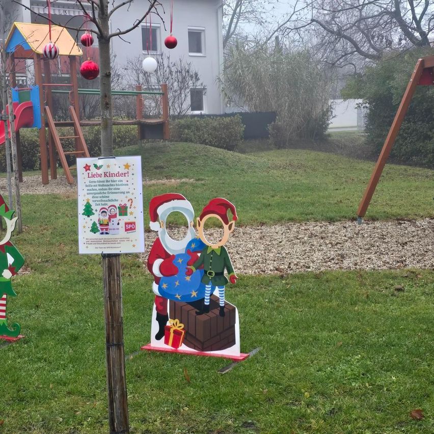 Eine weihnachtliche Ausschneidefigur mit Weihnachtsmann und Elfe steht in einem Park. Ein Schild ist in der Nähe, und Weihnachtsdekorationen schmücken die Bäume. Ein Haus und ein Spielplatz sind im Hintergrund.