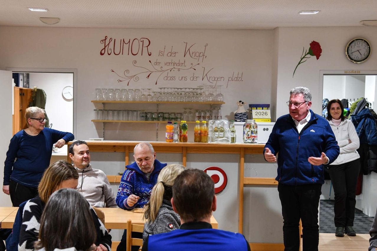 Ein Mann steht vor einer Gruppe von Menschen, die an Tischen sitzen, und spricht zu ihnen, während er ein Mikrofon hält. An der Wand hinter ihnen ist ein Zitat angebracht.