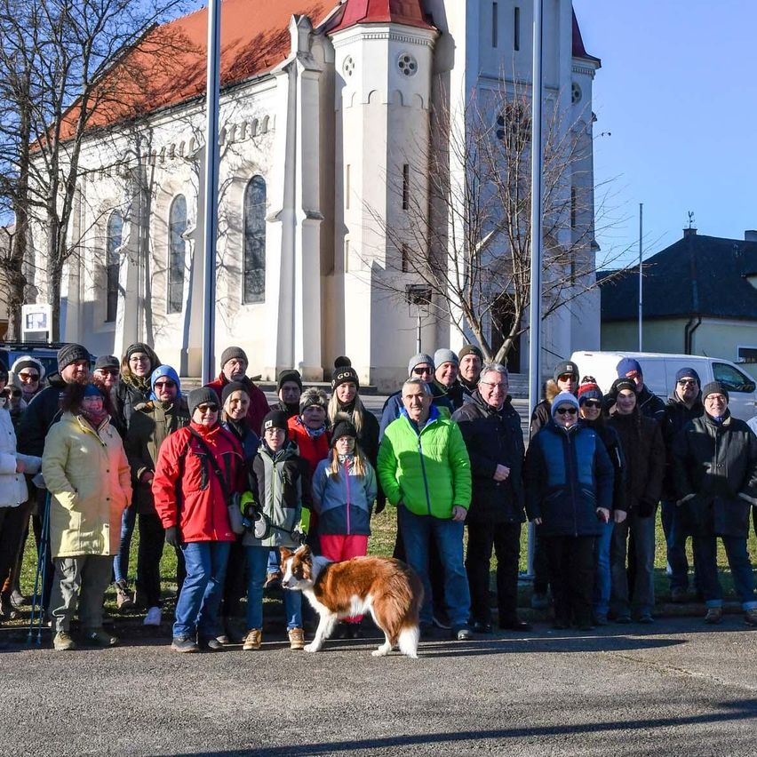 Eine Gruppe von Menschen steht draußen, einige in Winterkleidung, mit einem Hund davor. Sie stehen vor einer Kirche, mit einem Fahnenmast und Bäumen im Blick.