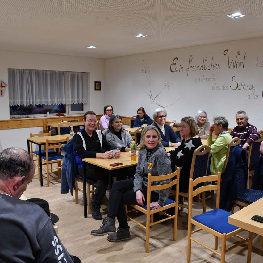 Eine Gruppe von Menschen sitzt um Tische in einem Restaurant mit einer Wandmalerei. Eine Frau in einer grauen Kapuzenjacke lächelt in die Kamera. Der Raum hat Holzböden und eine weiße Decke.