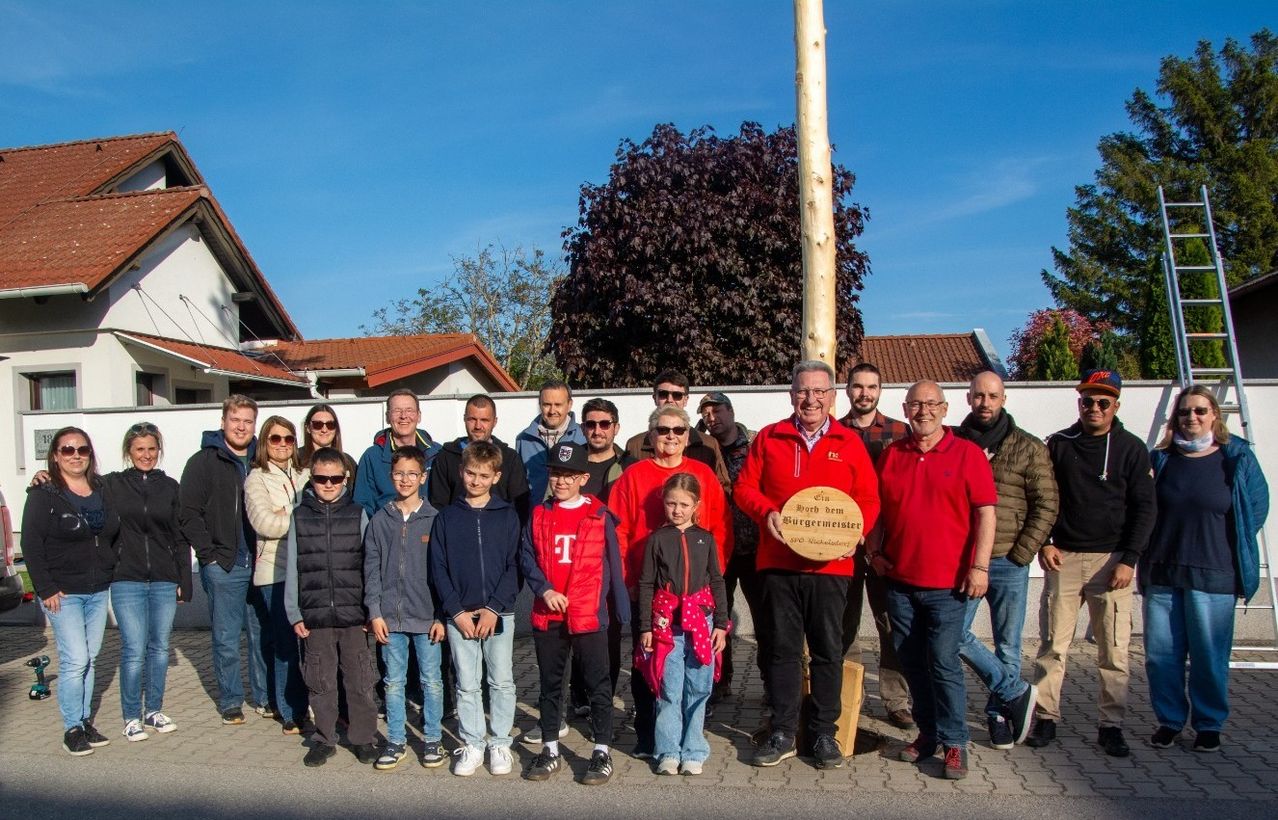 Eine Gruppe von Erwachsenen und Kindern posiert für ein Foto vor einer weißen Mauer. Ein Mann hält eine Plakette mit der Aufschrift 'Roth dem Burgermeister'.