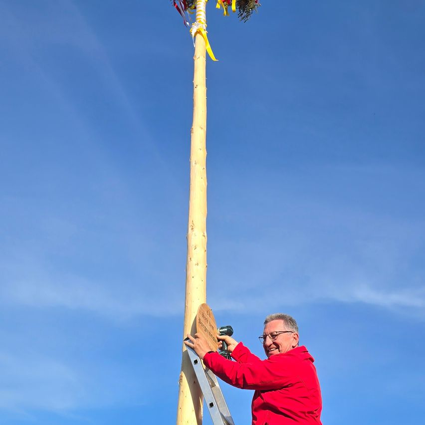 Ein Mann in einem roten Mantel klettert eine Leiter hinauf, um ein Schild an einen hohen Holzpfahl mit bunten Bändern zu befestigen. Der Himmel ist klar blau.