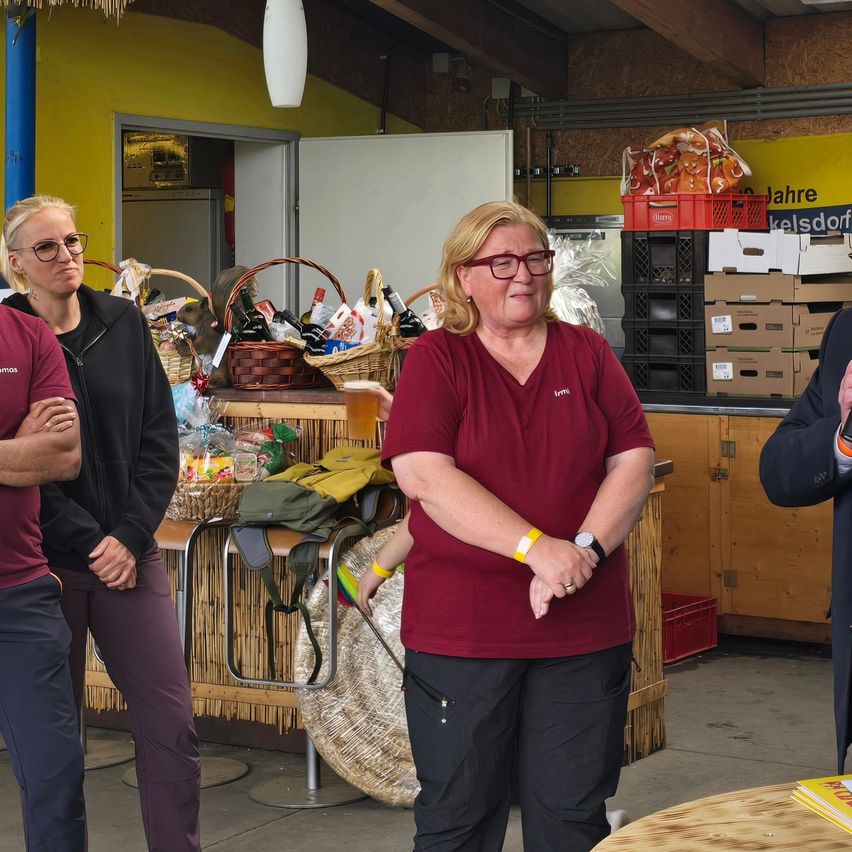 A group of people are standing in a warehouse, one woman is smiling while the others look serious. There are baskets and bottles on the table.