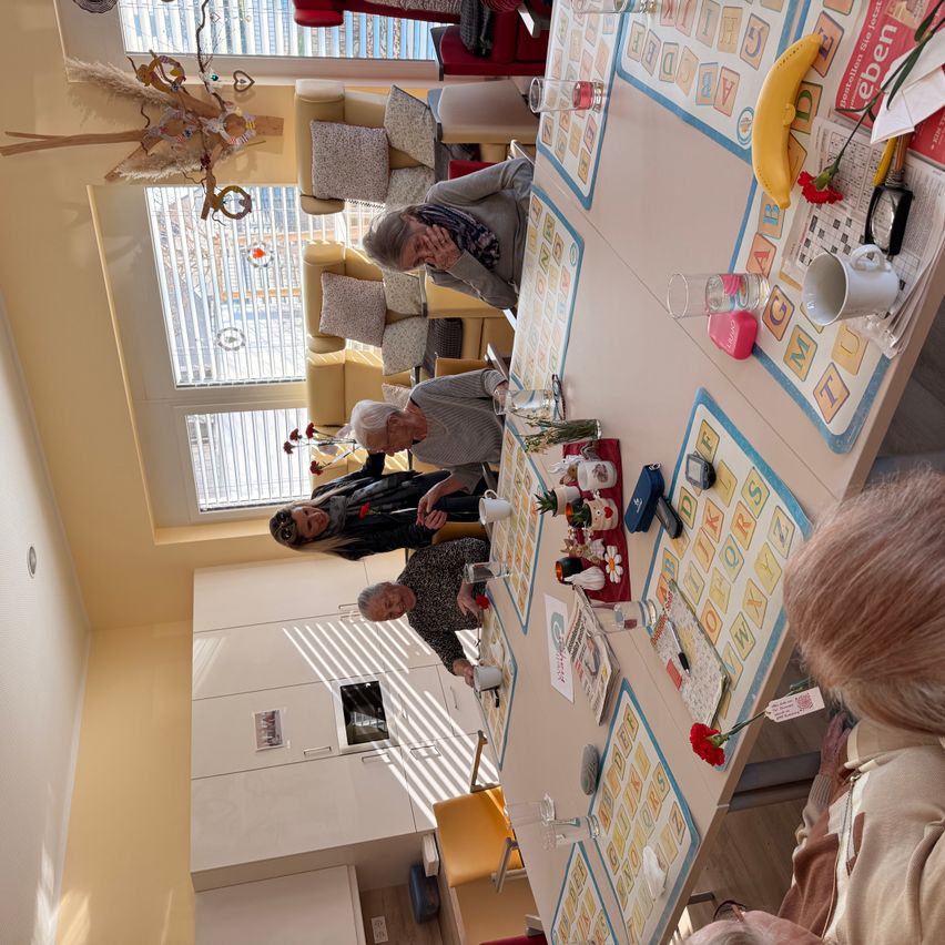 Fünf Personen sitzen um einen Tisch in einem Raum, möglicherweise spielen sie ein Brettspiel. Auf dem Tisch stehen Tassen, eine Banane und eine Blume.