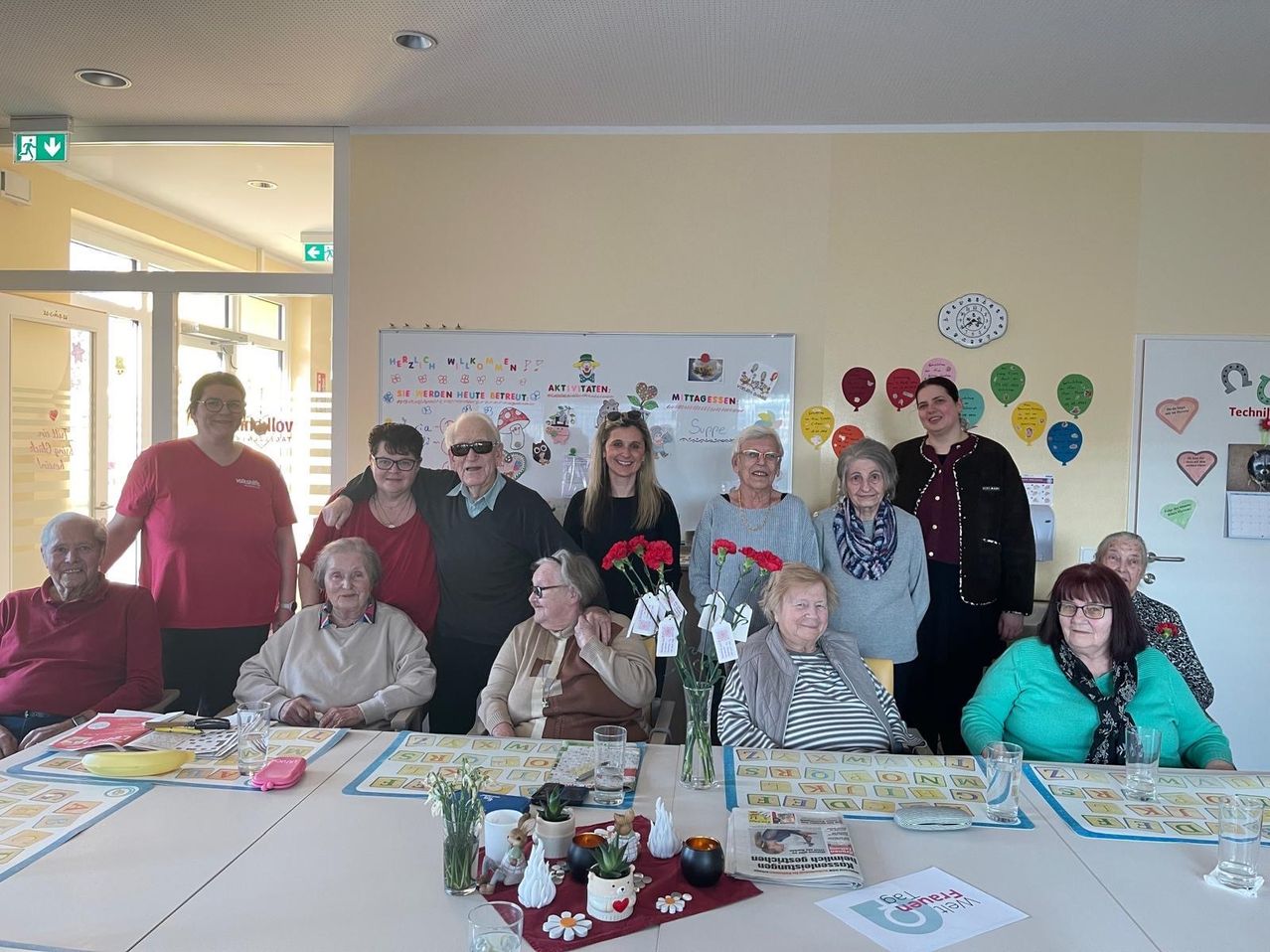 Eine Gruppe älterer Erwachsener steht vor einem Tisch mit Spielen, Luftballons und einer Uhr an der Wand. Der Raum hat eine gelbe Wand und eine Glastür.