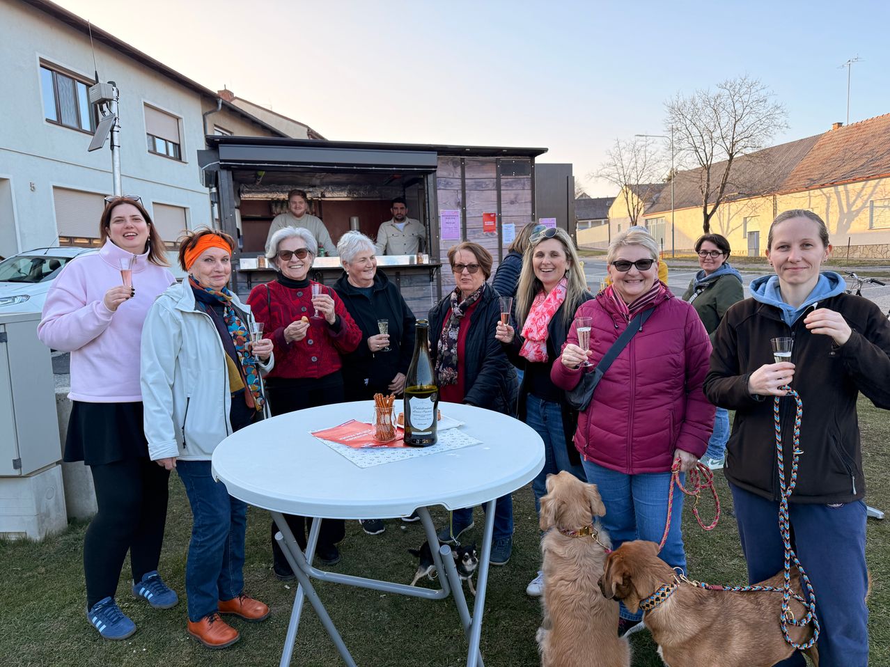 Eine Gruppe lächelnder Frauen steht um einen Tisch mit Weingläsern und zwei Hunden an der Leine. Dahinter steht ein offener Imbisswagen, in dem sich Menschen befinden. Der Himmel ist klar.