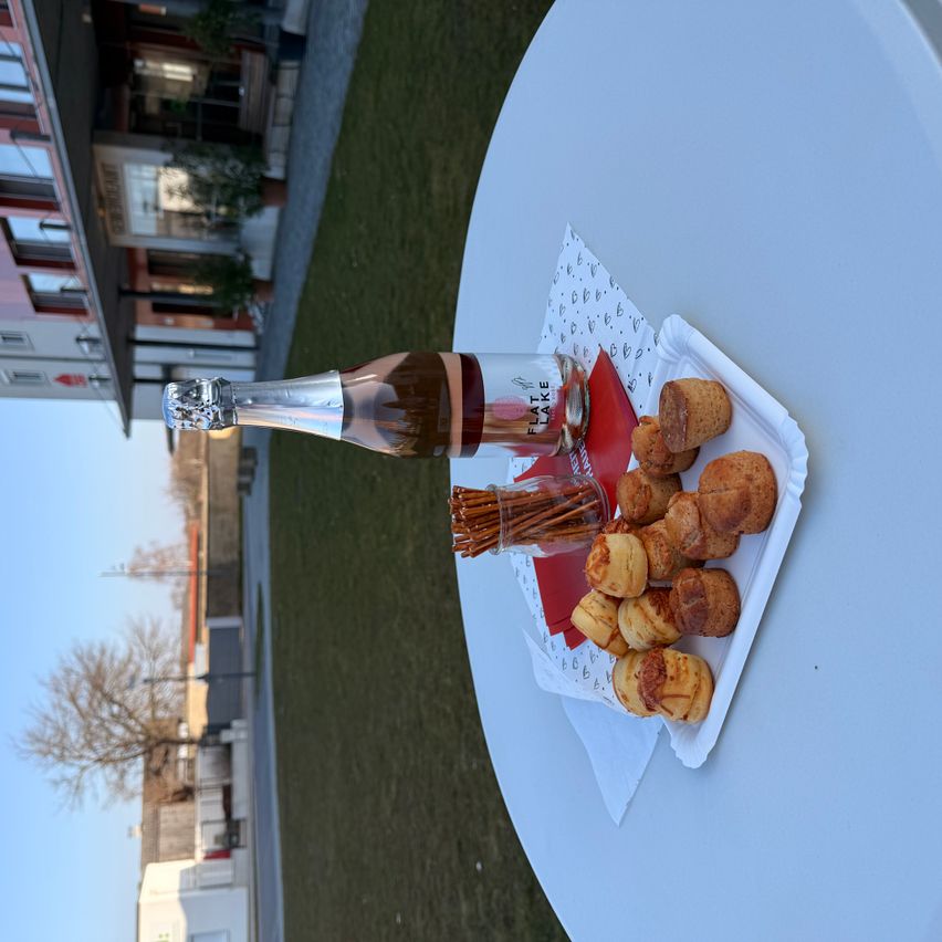 Eine Flasche Sekt, Brezeln und Mini-Quiches auf einem weißen Tisch. Der Tisch ist von einer Rasenfläche umgeben, im Hintergrund steht ein Gebäude.