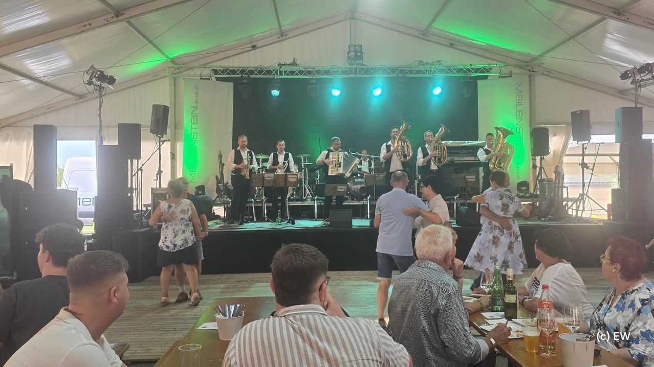 Eine Brass-Band spielt auf einer Bühne unter grünen Lichtern in einem Zelt, während einige Zuschauer zuschauen und tanzen.