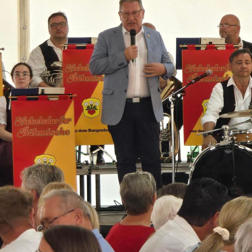 Ein Mann hält eine Rede auf der Bühne mit einem Mikrofon, während eine Band hinter ihm spielt. Das Publikum sitzt vor der Bühne.