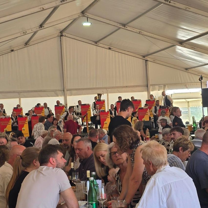 Eine große Gruppe von Menschen ist in einem Zelt versammelt, einige sitzen, andere stehen. Eine Band spielt auf der Bühne mit roten Bannern. Das Publikum ist engagiert, einige trinken aus Gläsern.