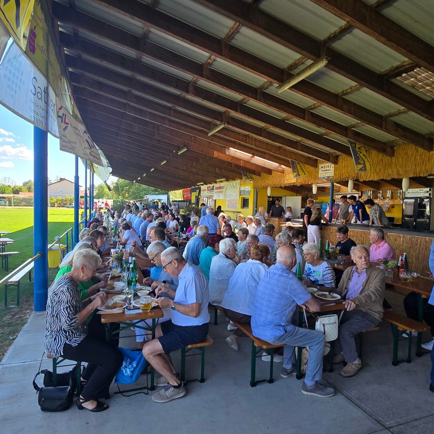 Eine große Gruppe von Menschen speisen im Freien unter einem Pavillon mit Holzbalken und einer gelben Wand. Sie sitzen an Tischen mit Essen und Getränken.