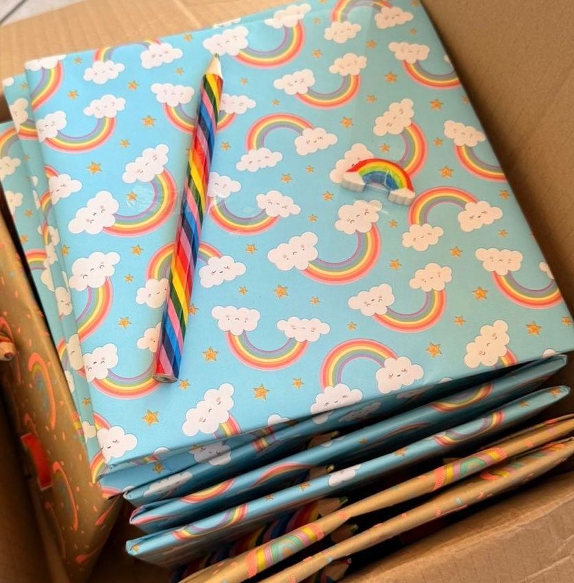 Ein Stapel bunter Notizbücher mit Regenbogen- und Wolkendesigns, einem Stift und einer Regenbogen-Papierklammer obenauf.