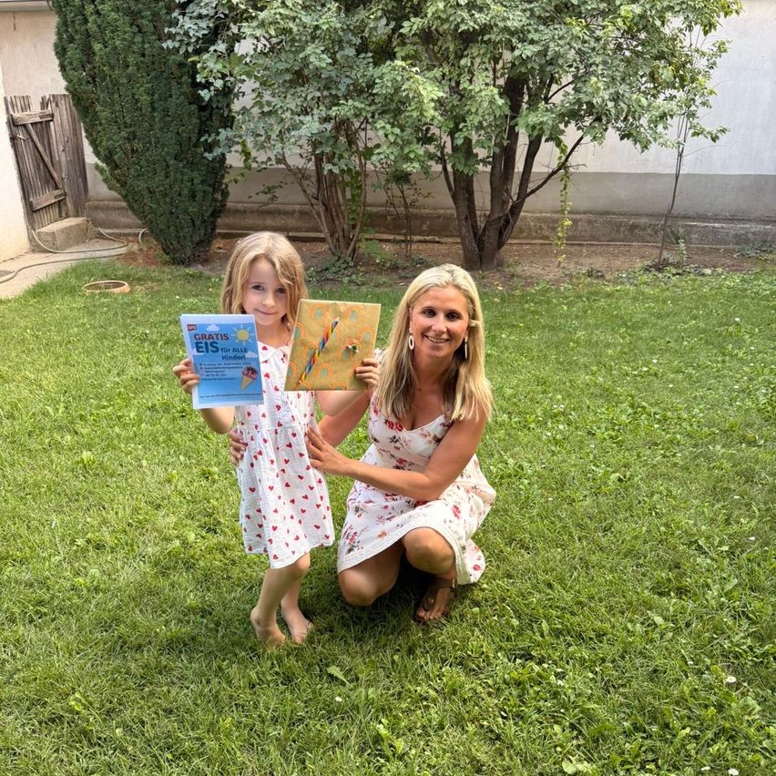 Eine Frau und ein junges Mädchen knien auf einem Rasen, lächeln und halten Bücher. Das Mädchen hält ein Buch mit dem Titel 'Gratis EIS für Alle Kinder'. Hinter ihnen sind Bäume und ein Gebäude.