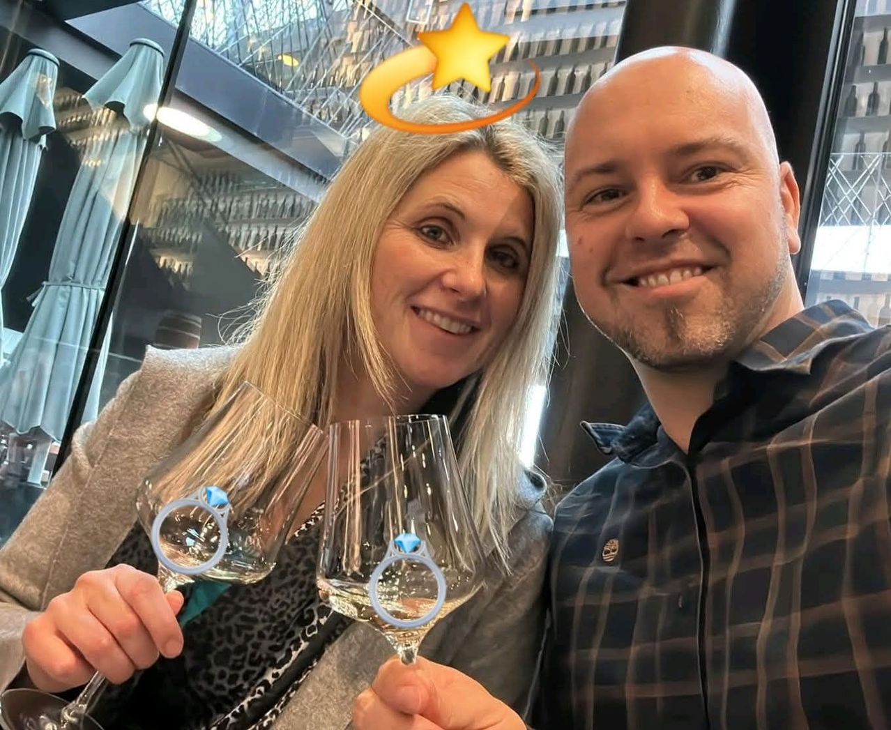 A man and woman hold wine glasses with rings in them, smiling for a photo inside a building.