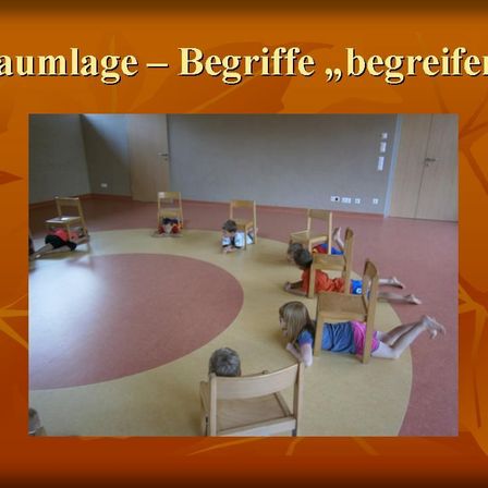 Eine Gruppe von Kindern liegt auf dem Boden in einem Kreis, wahrscheinlich in einem Klassenzimmer. Der Raum hat Holztüren und eine weiße Wand. Der Boden ist mit einem roten Teppich bedeckt.