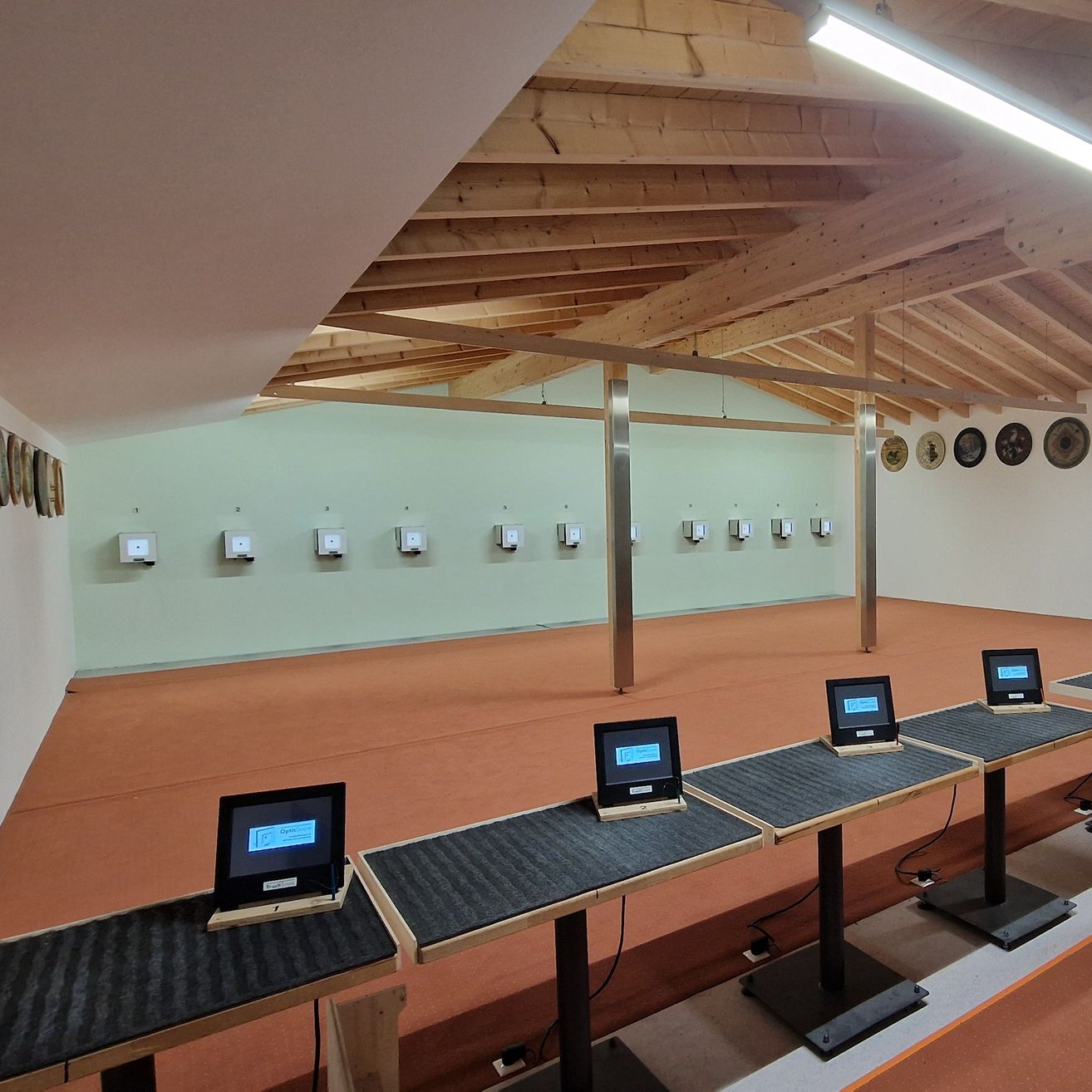 Eine überdachte Schießanlage mit Holzbalken an der Decke und elektronischen Monitoren auf Tischen. Mehrere Ziele sind an der Wand befestigt.