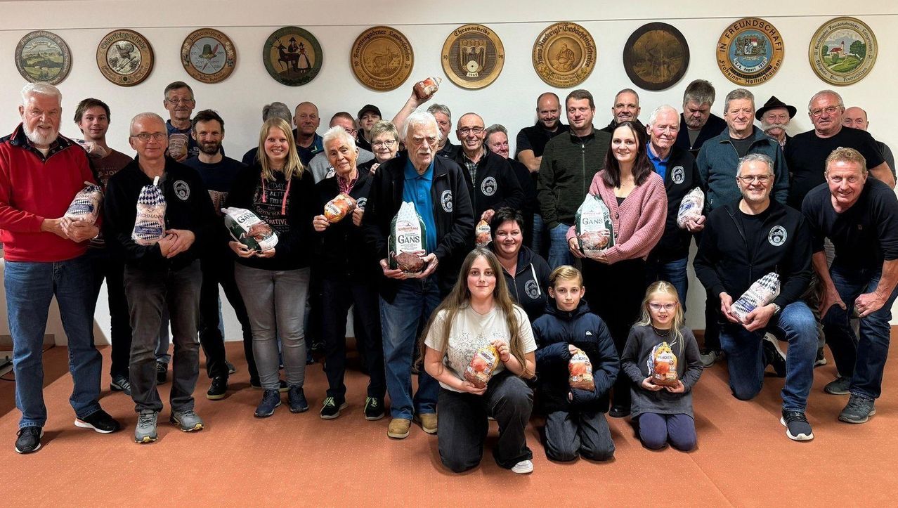 A group of people, some standing and some sitting, are posing for a picture. They are holding bags of meat. They are standing in front of a wall with several round objects attached to it.
