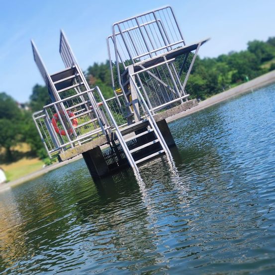 Eine Metallplattform mit Treppen und Handläufen ragt in einen See mit einem roten Rettungsring an der Seite. Bäume und ein klarer Himmel sind im Hintergrund.