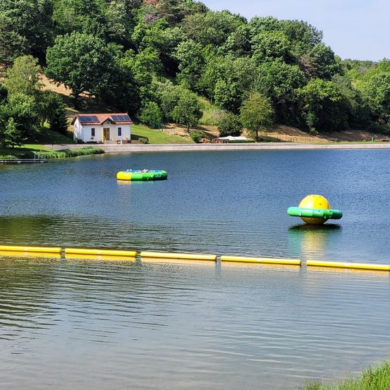 Eine friedliche Seelandschaft mit einem kleinen, weißen Haus mit Solarpanelen auf einem Hügel, umgeben von üppigem Grün. Zwei aufblasbare Schwimmkörper sind auf dem Wasser, einer gelb und grün, der andere grün und gelb.