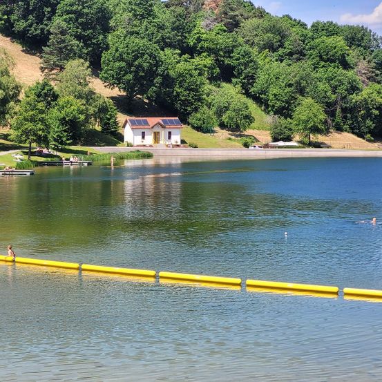 Ein ruhiger See mit einem kleinen Haus am Ufer. Bäume und Berge im Hintergrund. Einige Leute schwimmen im Wasser.