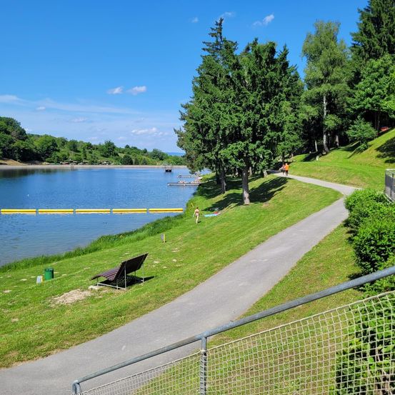 Ein ruhiger See mit einem grasbewachsenen Ufer, einem Gehweg und einer Bank unter blauem Himmel. Bäume und Büsche säumen den Pfad, und eine Person in Orange steht in der Nähe.