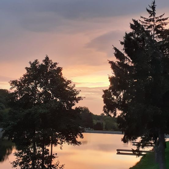 Ein ruhiger Seeblick bei Sonnenuntergang, mit einem See, der die warmen Farben des Himmels reflektiert. Bäume säumen das Ufer, und ein Steg erstreckt sich ins Wasser.