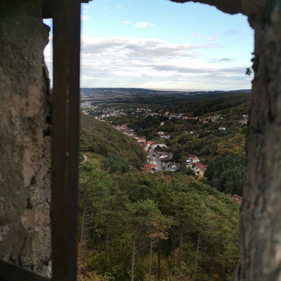 Ein Blick aus einer Steinmauer zeigt ein Dorf, umgeben von Bäumen, unter einem bewölkten Himmel mit Bergen in der Ferne.