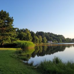 Ein ruhiger See, umgeben von üppigem Grün und Bäumen. Ein kleines Haus steht am Ufer. Der Himmel ist klar und blau.