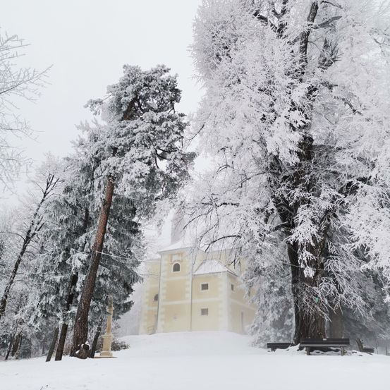 Eine verschneite Landschaft mit einer Kirche und mit Schnee bedeckten Bänken im Vordergrund. Die Kirche hat ein weißes Dach und ist von schneebedeckten Bäumen umgeben.
